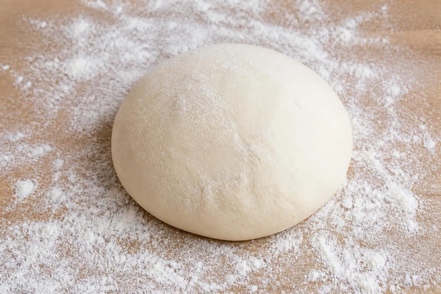 une boule de pâte à pizza crue posée sur un plan de travail fariné, avec un peu de farine éparpillée autour